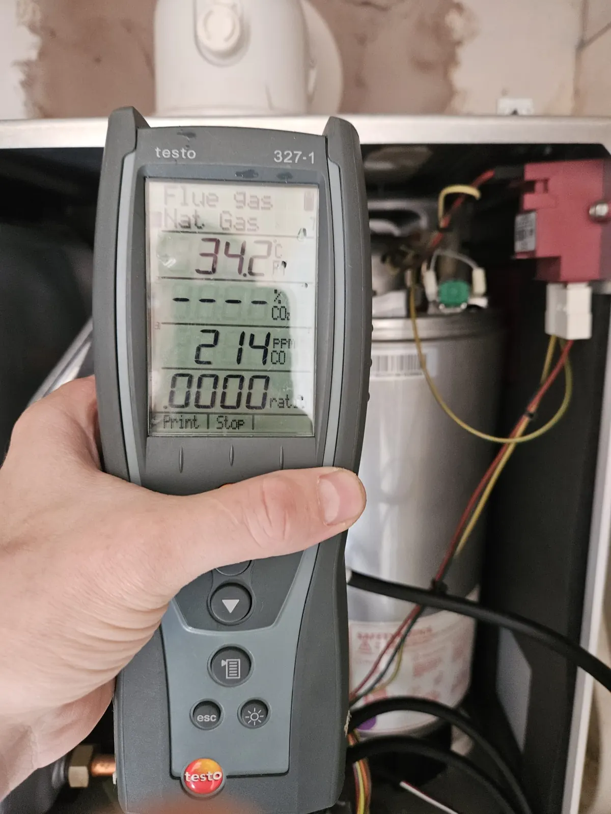 A hand holding a flue gas analyser in front of an open boiler, with a reading on the display